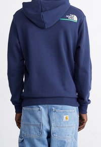 Mörkblå hoodie med dragsko i huvan, ribbade muddar och fåll. Har en vit logotyp på axeln och ett mjukt, strukturerat material.