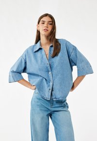 Lyseblå denim skjorte med en afslappet pasform, korte ærmer og knaplukning foran. Stoffet har en glat tekstur og et afslappet design.