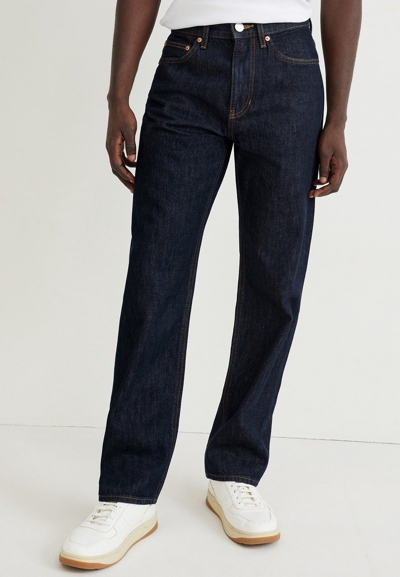 Man i mörkblå raka jeans, kombinerade med vita sneakers och en vit skjorta, som står mot en enkel ljus bakgrund.