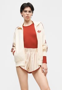 Νεαρό άτομο με διάφανα γυαλιά, κρεμ μπουφάν Adidas, κόκκινη μπλούζα με ριμπ ύφανση και κρεμ σορτς με κόκκινες κάθετες ρίγες, σε απλό φόντο.