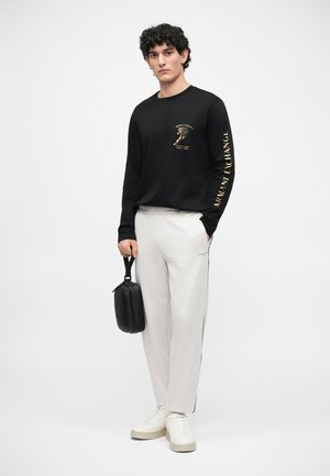 Maglietta nera a maniche lunghe con stampa grafica dorata, abbinata a pantaloni da tuta grigi chiari con strisce laterali. Il modello tiene una piccola borsa nera.