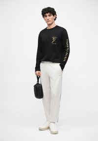 Melna krekla ar garām piedurknēm un zelta grafisko druku, apvienots ar gaiši pelēkiem sporta biksēm, kurām ir sānu svītras. Modelis tur nelielu melnu somiņu.