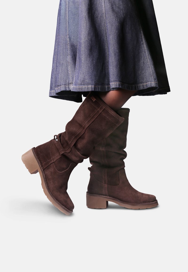 Bottes en daim marron mi-mollet avec de petits talons blocs, portées avec une jupe en jean bleu foncé, montrées jambes croisées sur fond blanc.
