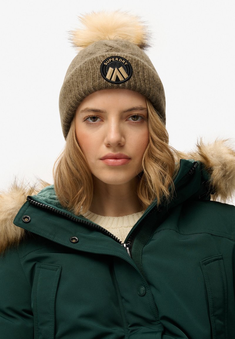 Gebreide muts in kaki kleur met een pluizige pompon, voorzien van een ronde zwarte patch met het "Superdry" logo. Gecombineerd met een groene jas en een crème kleurige trui.
