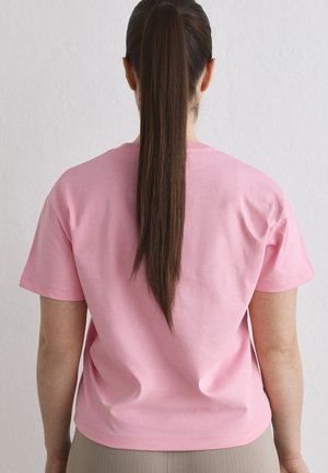 Femme aux longs cheveux noirs attachés en queue de cheval, portant un T-shirt rose ample, vue de dos sur un fond clair uni.