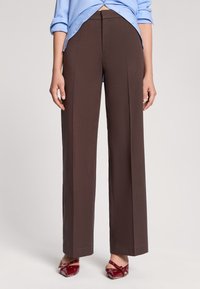 Pantaloni marroni a gamba larga con una texture liscia, vita alta e vestibilità sartoriale, abbinati a una camicia azzurro chiaro e scarpe rosse.