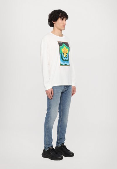 Witte longsleeve t-shirt met een kleurrijk grafisch ontwerp met groene, gele en blauwe golvende patronen en twee sterren. Gepaard met blauwe jeans.