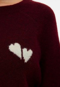 Maglione borgogna con una morbida texture lavorata a maglia, presenta due motivi a cuore bianchi nella zona del petto. Design classico con collo rotondo.