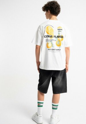 Osoba nosząca białą koszulkę z grafiką owoców cytrusowych i napisem, czarne krótkie spodenki do kolan, białe trampki oraz białe skarpetki z zielonymi paskami.