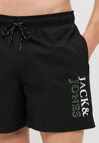 Fekete úszónadrág, gumis derekkal és zsinórral, oldalsó zsebekkel, valamint fehér és zöld "JACK & JONES" felirattal az oldalon.