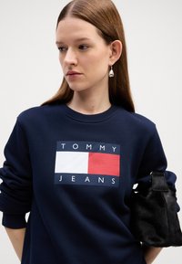 Felpa blu navy con un logo rettangolare che presenta il testo "TOMMY JEANS" e una grafica rossa e bianca. Borsa a tracolla in pelliccia nera.