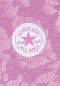 Patch rond blanc de Converse All Star avec une étoile rose et du texte sur un fond en tissu texturé rose.