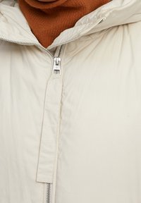 Veste rembourrée blanche avec un col haut, dotée d'une fermeture éclair argentée et d'un tissu texturé. Un pull en tricot marron est visible en dessous.