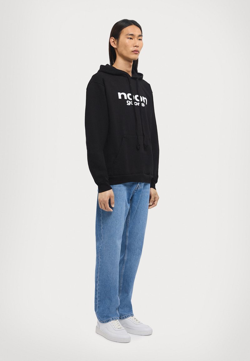 Schwarzer Hoodie mit weißem "noon"-Logo, Kängurutasche, Kapuze mit Kordelzug; kombiniert mit lässigen blauen Jeans und weißen Sneakers.