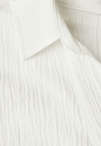 Tela blanca texturizada con un sutil patrón arrugado. Presenta un cuello con solapa y un material suave y ligero.