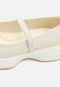Chaussure Mary Jane couleur crème avec bride en tissu, semelle intérieure rembourrée, semelle blanche épaisse et subtil accent doré proche du bord de la semelle.