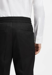 Schwarze Hose mit elastischem Bund, ausgestattet mit zwei seitlichen Taschen, dazu ein teilweise sichtbares, weißes Langarmshirt mit Struktur.