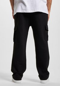 Pantaloni de trening negri, cu două buzunare laterale, dintr-un material moale, având o croială lejeră și o textură netedă. Pantofi albi vizibili în partea de jos.