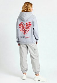 Felpa grigia con un grande motivo a cuore rosso sul retro, presenta il testo "SWEET heart", abbinata a pantaloni cargo grigi chiari e sneakers bianche.