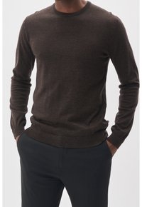 Man i en åtsittande brun långärmad tröja som är lätt instoppad i svarta byxor med händerna i fickorna mot en enkel bakgrund.