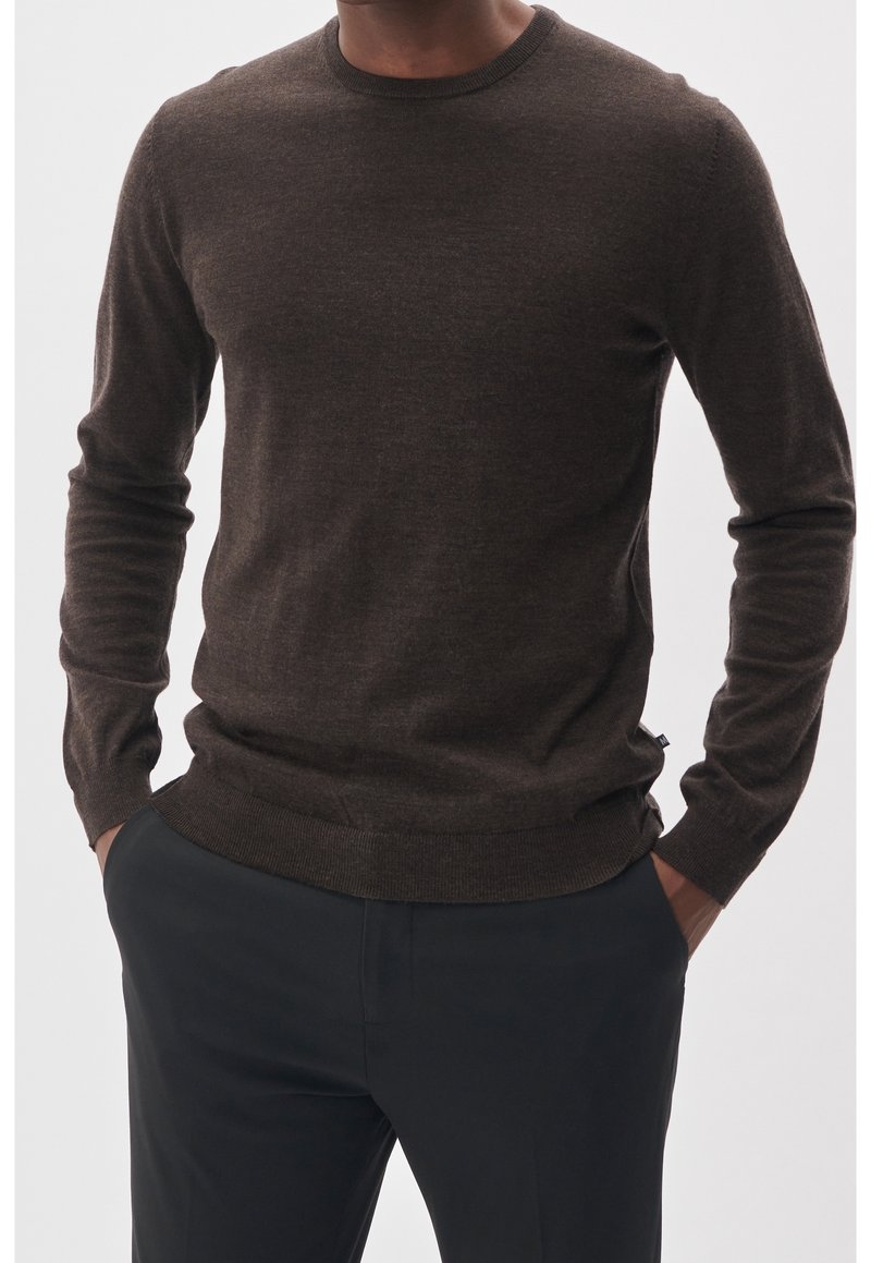 Man i en åtsittande brun långärmad tröja som är lätt instoppad i svarta byxor med händerna i fickorna mot en enkel bakgrund.