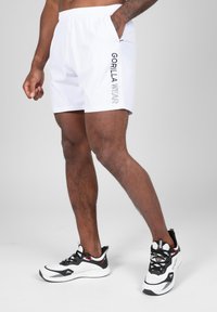 Witte shorts met een elastische tailleband en zijzakken, met "GORILLA WEAR" in zwarte tekst aan de linkerkant. Zwart-witte sportchoenen.