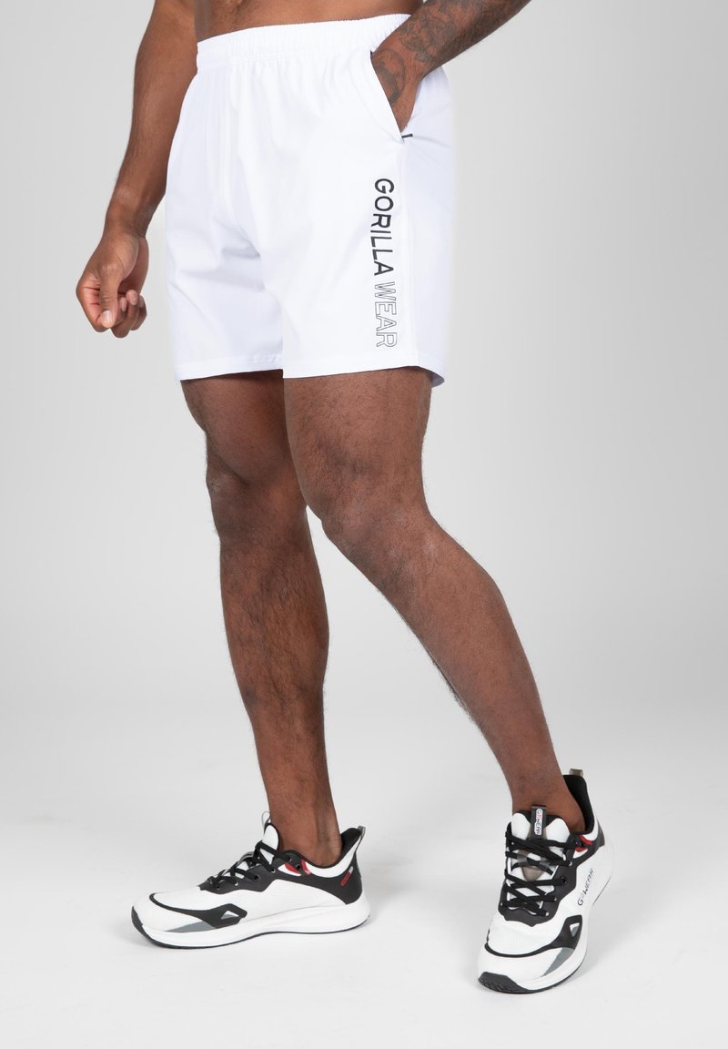 Witte shorts met een elastische tailleband en zijzakken, met "GORILLA WEAR" in zwarte tekst aan de linkerkant. Zwart-witte sportchoenen.
