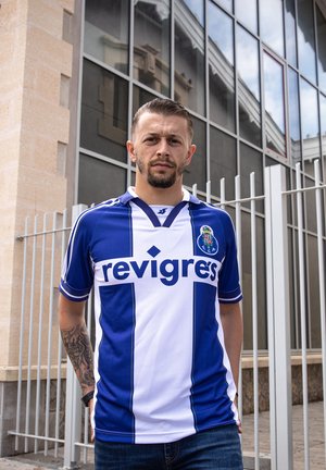 Un homme avec des cheveux courts et une barbe porte un maillot de football bleu et blanc du FC Porto, se tenant à l'extérieur près d'une clôture en métal et d'un bâtiment en verre.
