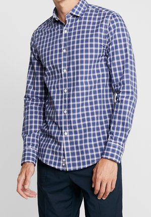 Mann trägt ein langärmeliges Hemd mit blau-weiß kariertem Muster und dunkle Hose vor schlichtem Hintergrund.