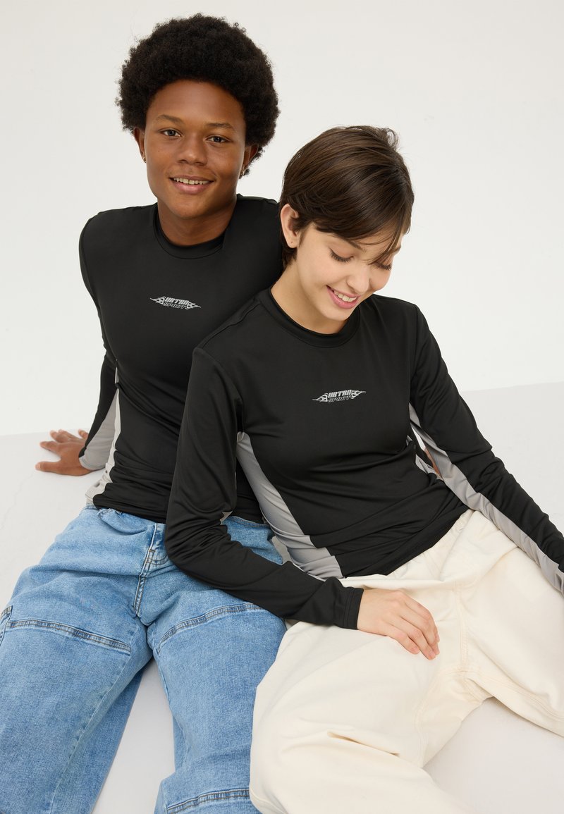 Sorte langermede rash guards har grå sidestykker. Personene har på seg casual denim og kremfargede bukser, og sitter mot en nøytral bakgrunn.