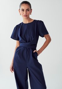 Temno modra kombinezonska obleka iz teksturirane tkanine, s kratkimi nabranimi rokavi, z pasom, obrnjenim proti visoki postavi, in širokimi hlačami s stranskimi žepi.