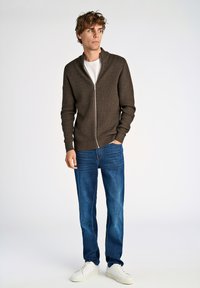 Maglione zip-up marrone con motivo testurizzato, abbinato a jeans blu e sneakers bianche. Il modello è in piedi davanti a uno sfondo neutro.