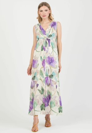 Žena v bílé maxi šaty bez rukávů s velkým fialovo-zeleným květinovým vzorem a béžové sandály na vysokém podpatku s pásky.