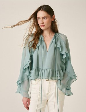 Mujer de cabello largo que lleva una blusa azul claro translúcida con volantes y pantalones blancos de talle alto, posando frente a un fondo liso.