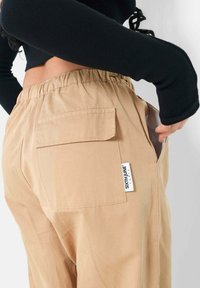 Tan cargo byxor med elastisk midjeband, sidoficka och bakficka med en sydd etikett. Slät tyg och avslappnad passform.