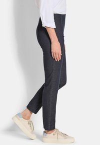 Vrouw in donkere slim-fit enkellange broek en beige veterschoenen, zijwaarts staand tegen een witte achtergrond.