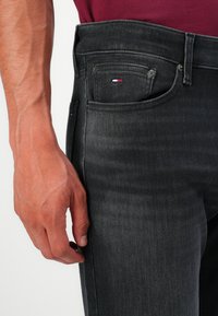 Männliche Hand, die nahe der Tasche von schwarzen Jeans aus Denim ruht, mit einem kleinen rot, weiß und blauen Logo auf der Münztasche, gekleidet in einem burgunderfarbenen Oberteil.