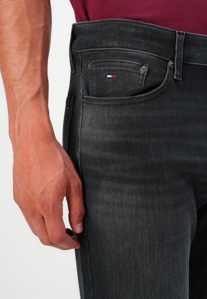 Male hand resting near the pocket of black denim jeans with a small red, white, and blue logo on the coin pocket, wearing a burgundy top.