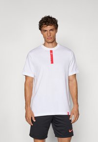 Hvid bomulds t-shirt med rød vertikal label på brystet. Korte ærmer, rund halsudskæring. Sammen med mørkegrå shorts. Simpelt design.