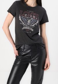 Kvinde iført sort grafisk t-shirt med ørn og teksten "Lover", parret med skinnende sorte læderbukser, hånd hvilende på hoften.