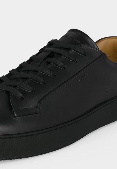 Μαύρο δερμάτινο αθλητικό παπούτσι με μαύρα κορδόνια, στρογγυλή μύτη και ορατή ραφή, που δείχνει μέρος της ταμπά εσωτερικής επένδυσης.