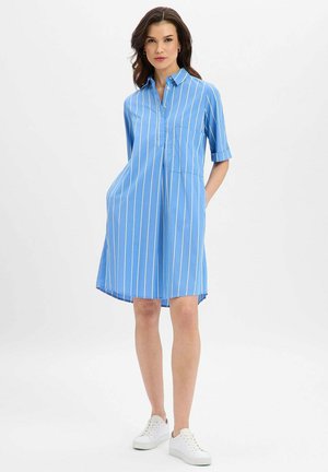 Frau trägt ein blaues, knielanges Kleid mit weißen vertikalen Streifen und weißen Turnschuhen, steht mit den Händen in den Taschen vor einem einfarbigen Hintergrund.