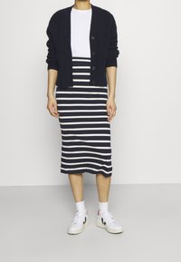 Kvinna klädd i en marinblå cardigan, vit t-shirt, marinblå och vit horisontellt randig midiklänning, vita strumpor och vita sneakers med svarta och röda detaljer.