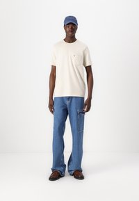 Light beige short-sleeve t-shirt with a left chest pocket, paired with blue denim cargo pants featuring side pockets, and brown shoes.