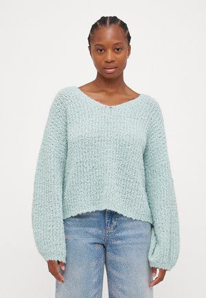 Pull en tricot bleu clair avec un col en V, des manches larges et un tissu texturé. Associé à un jean bleu délavé, créant un look décontracté.