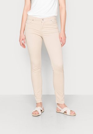 Kvinde iført beige tætsiddende bukser og hvide sandaler med krydsede stropper, står mod en ensfarvet hvid baggrund.