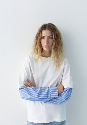 Fiatal nő hullámos szőke hajjal, fehér inget visel, amely alatt egy kék-fehér csíkos hosszú ujjú van, karját összefonva áll.