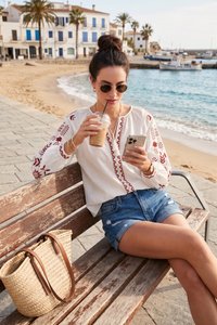 Femme en lunettes de soleil et blouse brodée, assise sur un banc au bord de la mer, buvant un café glacé et regardant son smartphone.