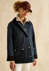 Jachetă impermeabilă de marină cu guler din catifea groasă maro, nasturi aurii și două buzunare frontale, suprapusă peste o cămașă în dungi. Capac maro cu logo.