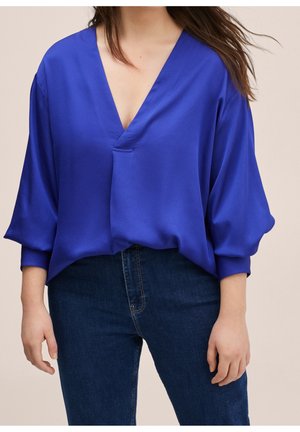 Kvinde iført en løstsiddende, dybblå bluse med V-hals og 3/4 ærmer, kombineret med mørkeblå jeans.